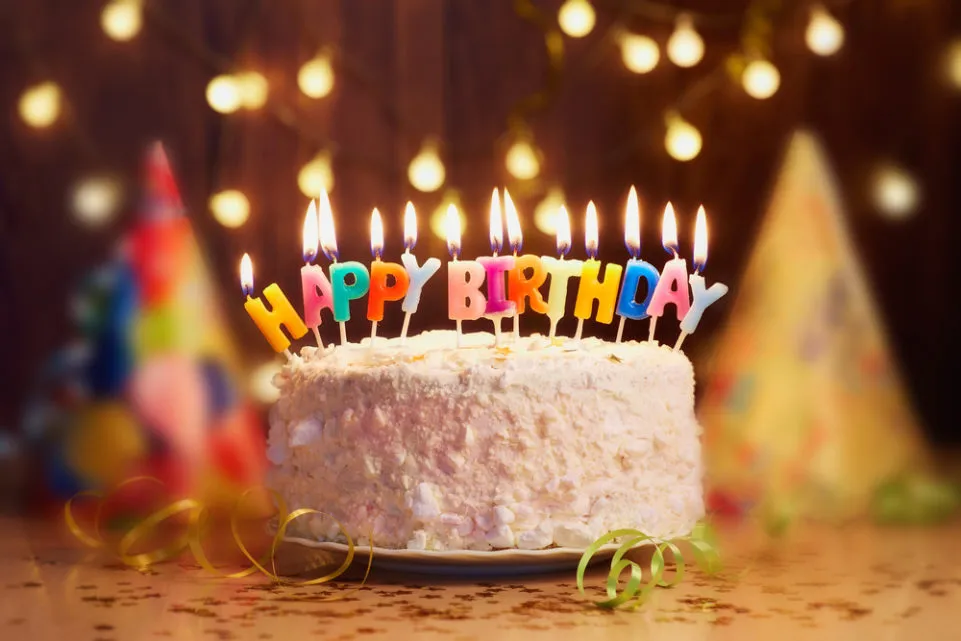 Bánh sinh nhật phủ kem trắng với hàng nến chữ nhiều màu ghép thành "HAPPY BIRTHDAY", phía sau là mũ tiệc và dây đèn mờ.